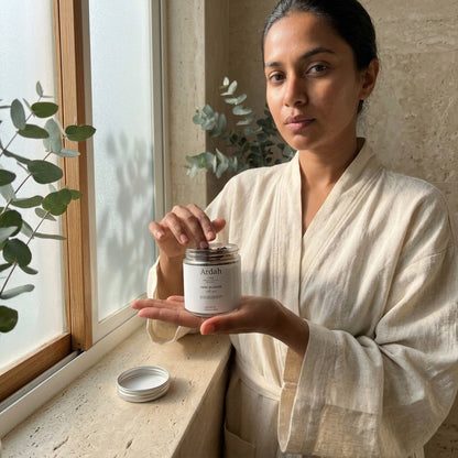 Woman in a white robe holding a jar labeled 'Anah' by a window with plants.